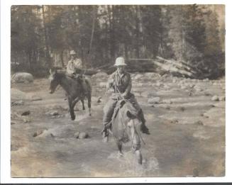Irv Lozier and Louise Rungius fording a stream on horseback.